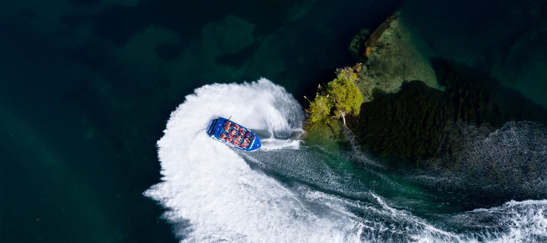 The image shows an aerial view of a blue jet boat labeled "hukafalls JET" making a sharp turn in the water, creating a large, white wake behind it. The boat is filled with passengers wearing life jackets as it navigates near a small, rocky outcrop covered